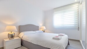 Una cama bien hecha con sábanas blancas, dos lámparas de noche, un cabecero gris y una ventana con persianas.