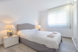 Una cama bien hecha con sábanas blancas, dos lámparas de noche, un cabecero gris y una ventana con persianas.