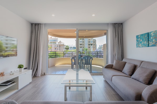 A modern living room with a glass table, sofa, and a balcony with a view of buildings.