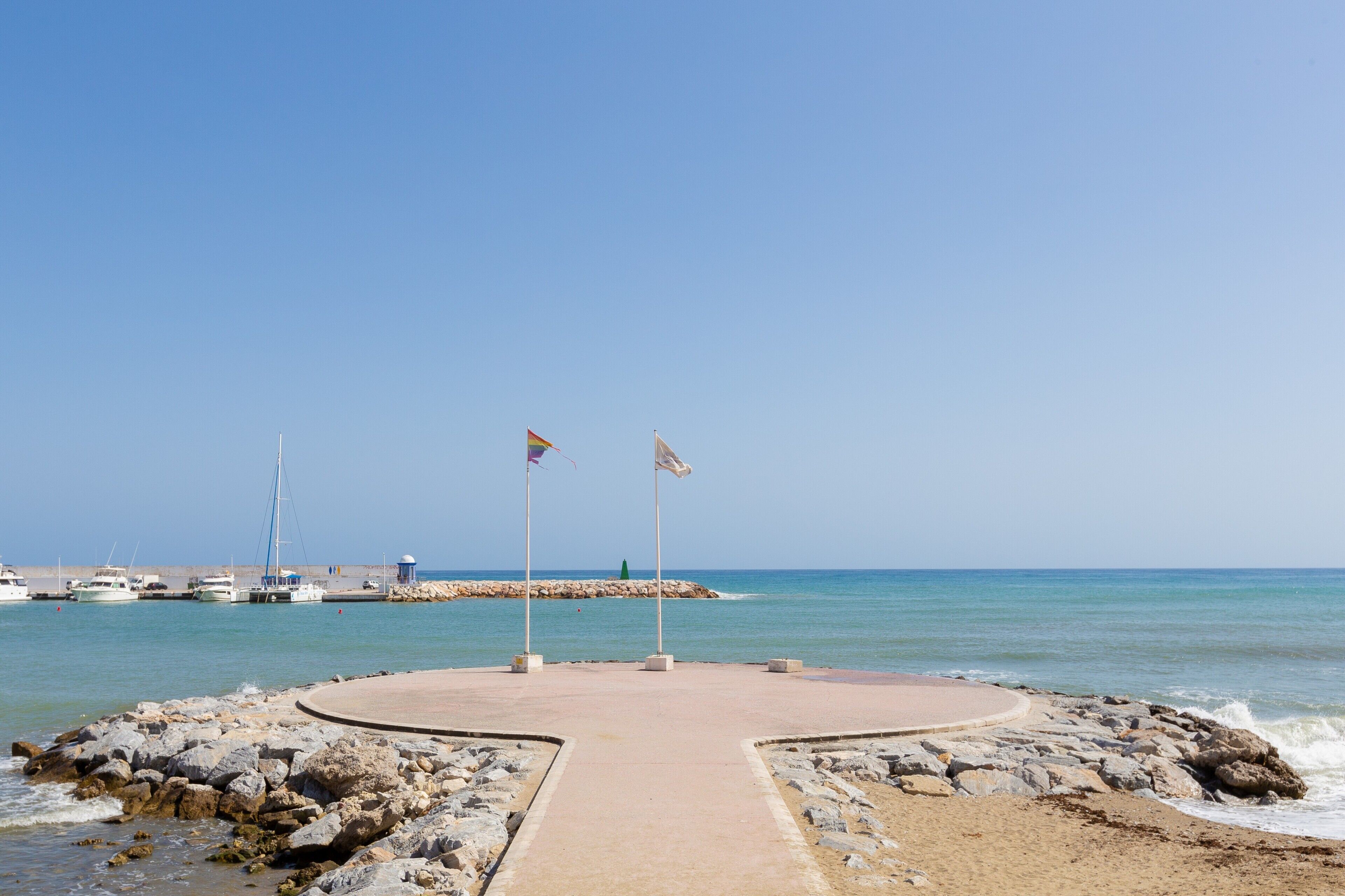 Ein kreisförmiger Steinweg führt zu einem Strand mit Flaggen, einer felsigen Küste und Booten im Hintergrund.
