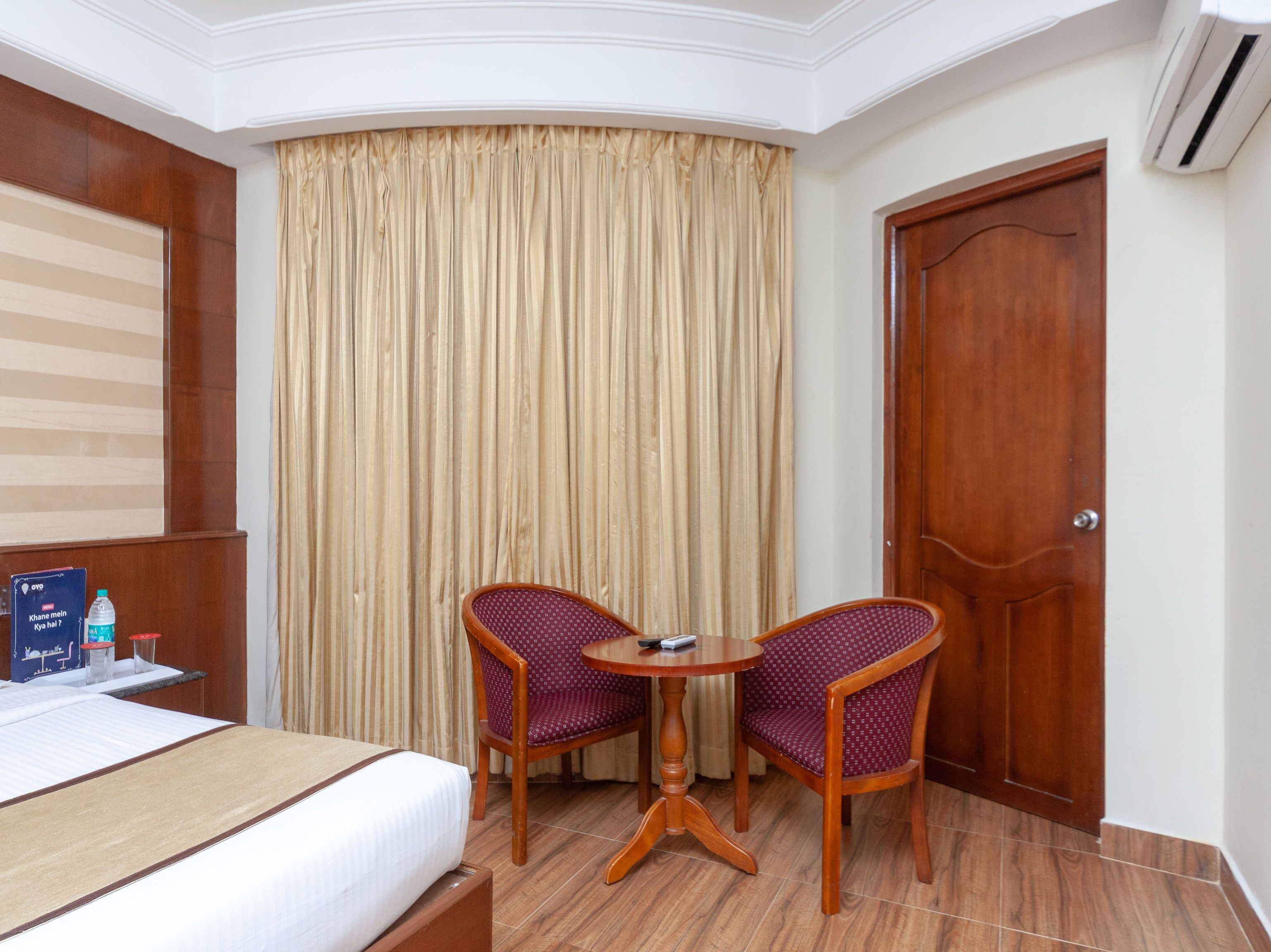 A hotel room with a bed, two chairs around a small table, a curtain, and a wooden door.