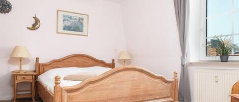 A bedroom with a wooden bed, two bedside tables with lamps, a window with a view, and a wall-mounted picture.