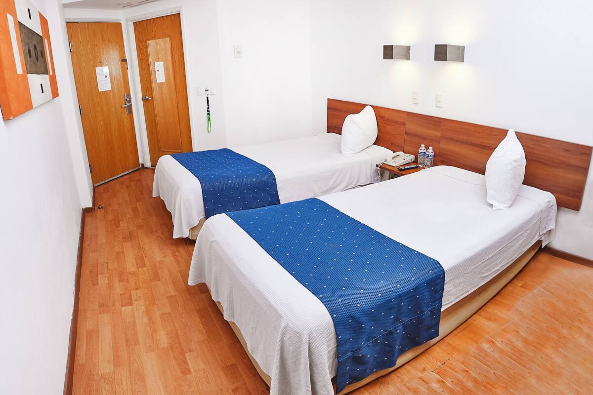 A hotel room with two beds, wooden flooring, a wooden headboard, a bedside table with a phone, and a wall-mounted light fixture.