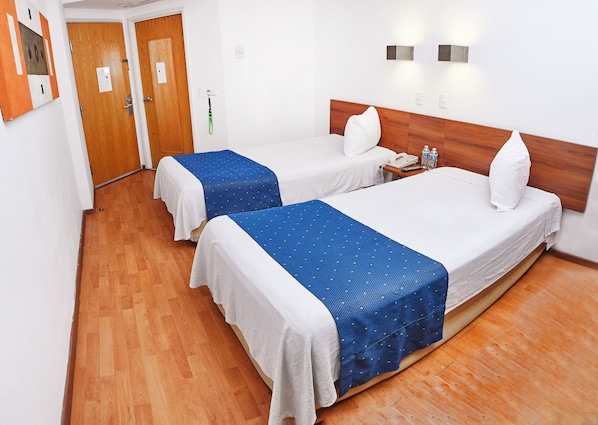 A hotel room with two beds, wooden flooring, a wooden headboard, a bedside table with a phone, and a wall-mounted light fixture.