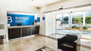 A business hotel reception area with a reception desk, a waiting area with a chair, and a view of the street through large windows. - BH Business Hotel Group (Reynosa)