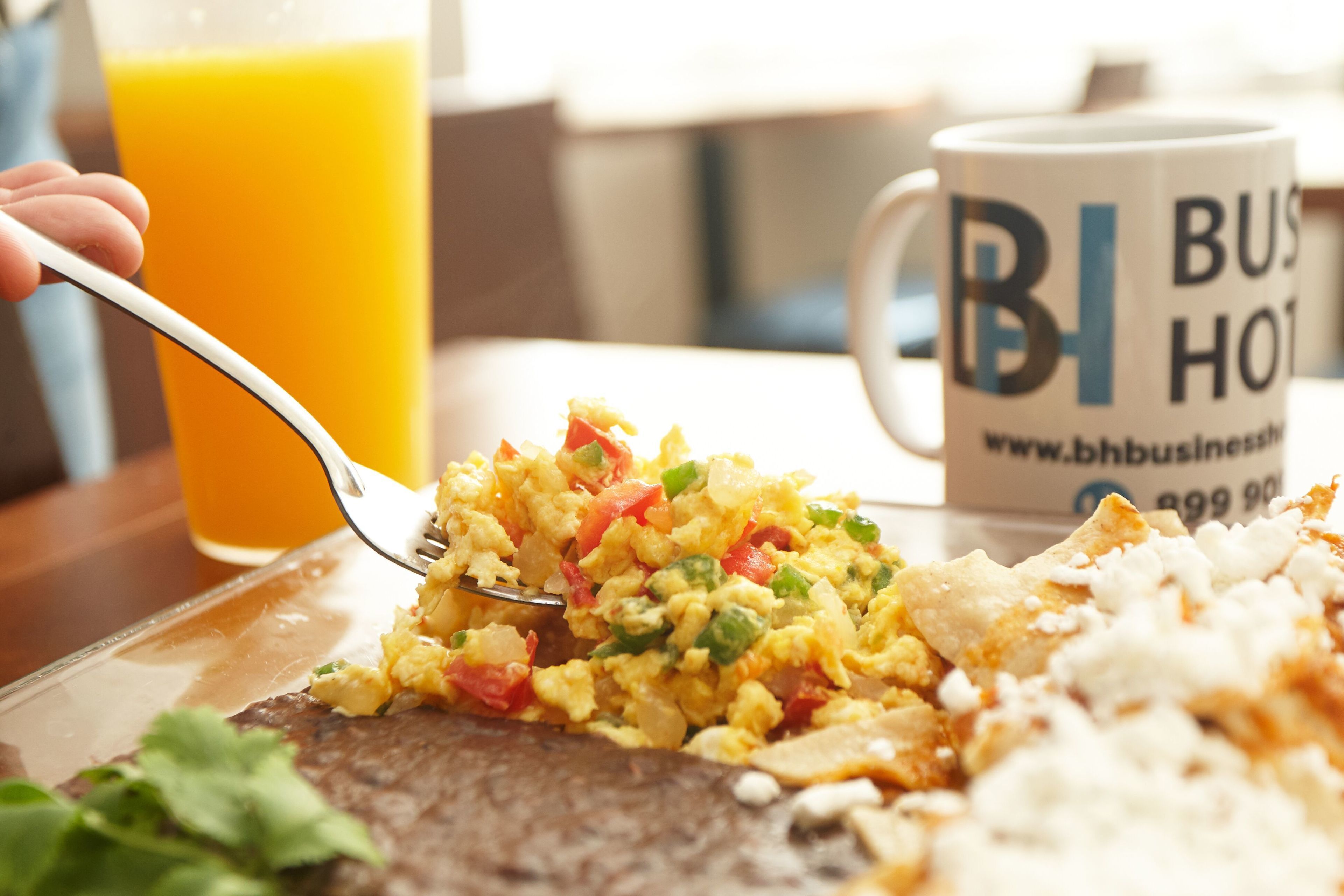 Un plato de desayuno con huevos revueltos, vegetales y tostadas, un vaso de jugo de naranja y una taza con el texto 'BH Business Hotel'.