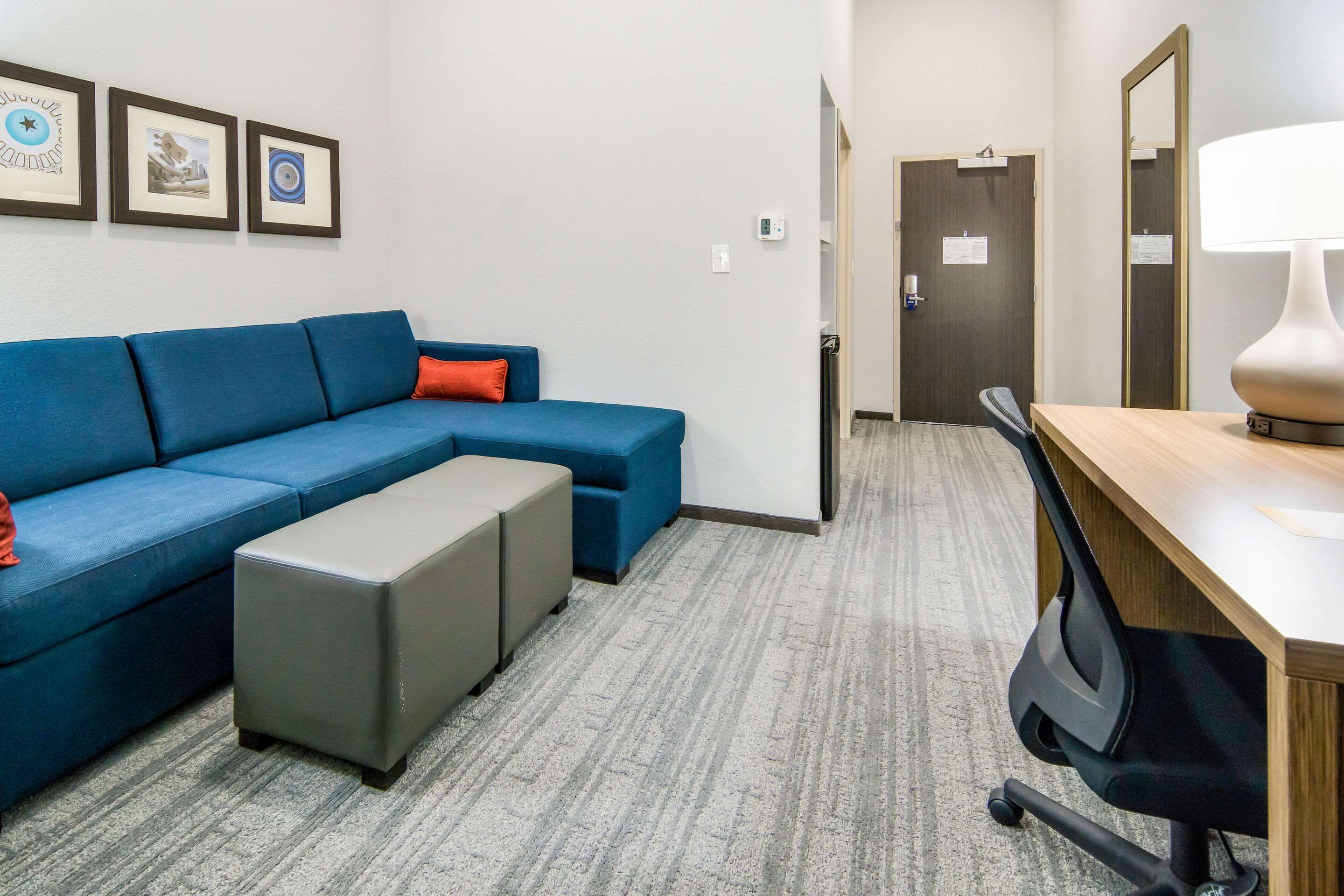 A hotel room with a blue sectional sofa, a grey ottoman, a wooden desk with a lamp, and a mirror. There are framed pictures on the wall and a door in the background.