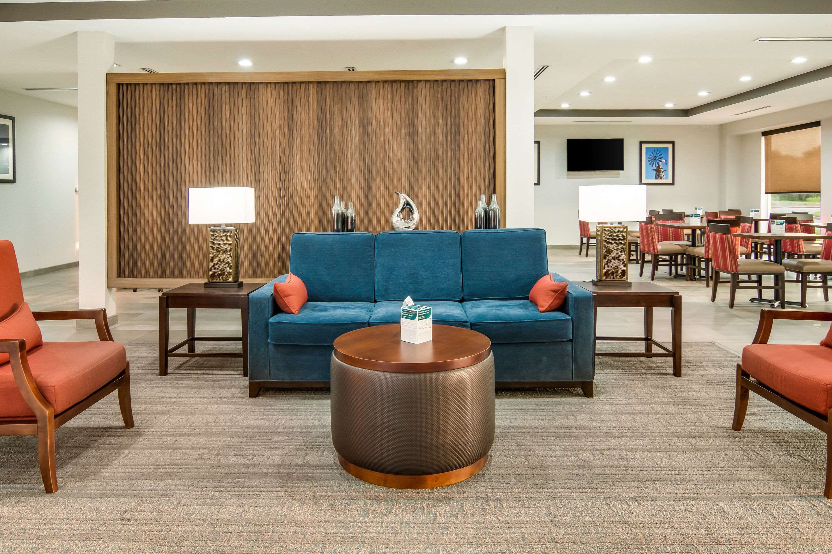 A modern lobby with a blue sofa, red chairs, a round table, and a wooden partition with a TV and artwork.
