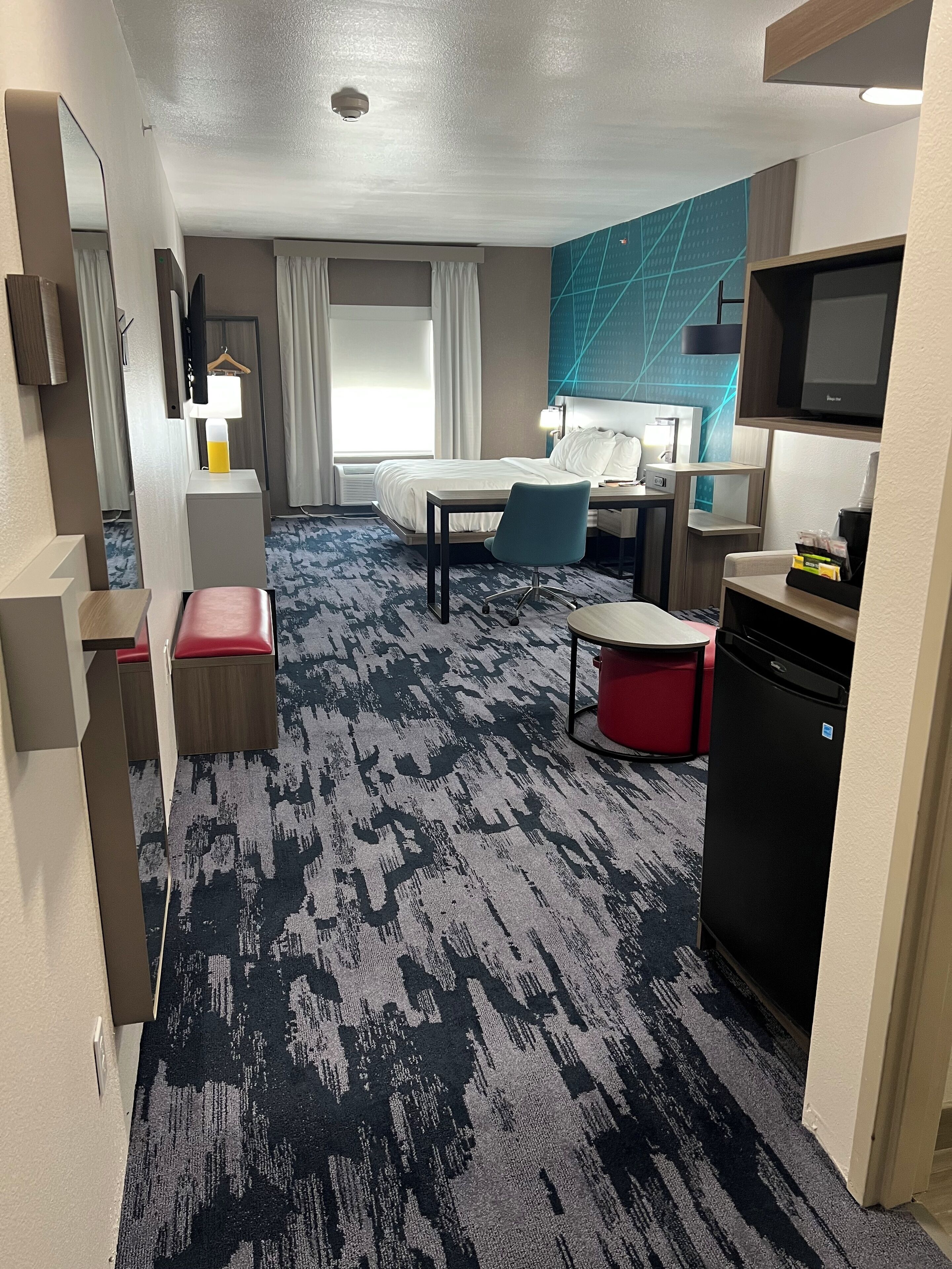 Quarto de hotel com cama, escrivaninha, cadeira, mesinha, um banco vermelho, televisão, mini-geladeira e micro-ondas.