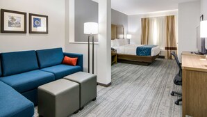 A hotel room with a blue sofa, a bed with a blue and white bedspread, a desk with a computer, and two framed pictures on the wall. - Comfort Suites DeSoto Dallas South (DeSoto)
