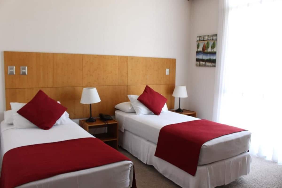 A hotel room with two beds, each with a red blanket and white bedding. There are bedside tables with lamps and a painting on the wall.