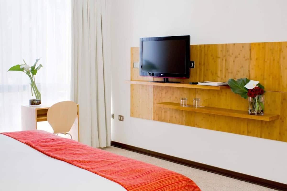 A hotel room with a bed, a TV mounted on a wooden panel, a desk with a chair, a vase with flowers, and a window with curtains.