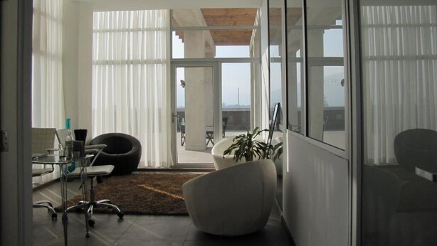 A modern interior with a glass partition, a desk with a chair, a large white chair, and a potted plant. - Open Hotel (Quillota)