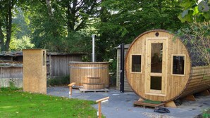 A wooden sauna structure with a door and windows, a small wooden cabin, and a hot tub in a wooded area. - Vacation home Friedrichstraße 16 (Laboe)