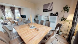 A modern living room with a large wooden dining table, comfortable seating, a flat-screen TV, and a sailboat painting on the wall. - Vacation home Friedrichstraße 16 (Laboe)