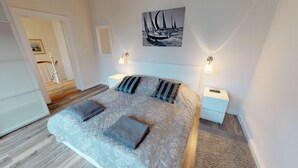 A bedroom with a bed, two bedside tables, a nightstand, a framed picture on the wall, and a door leading to another room. - Vacation home Friedrichstraße 16 (Laboe)