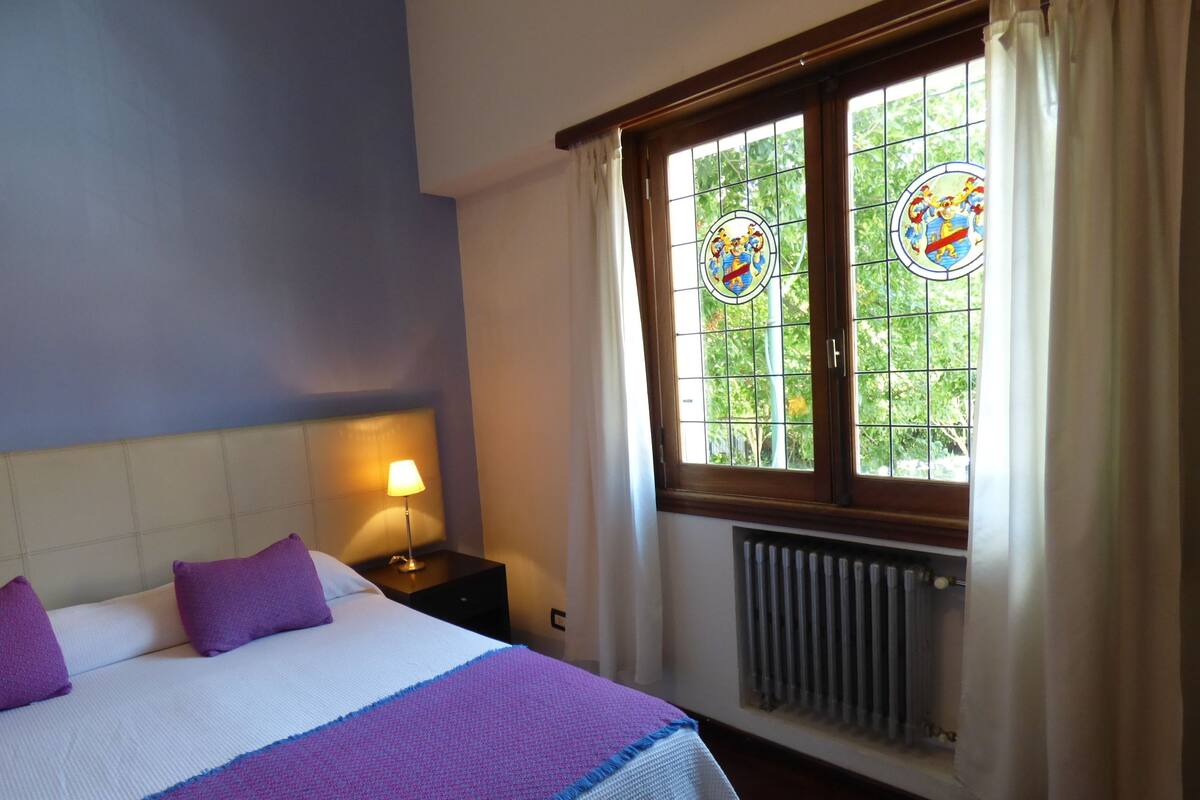 A hotel room with a bed, a nightstand with a lamp, a window with stained glass, and a radiator.