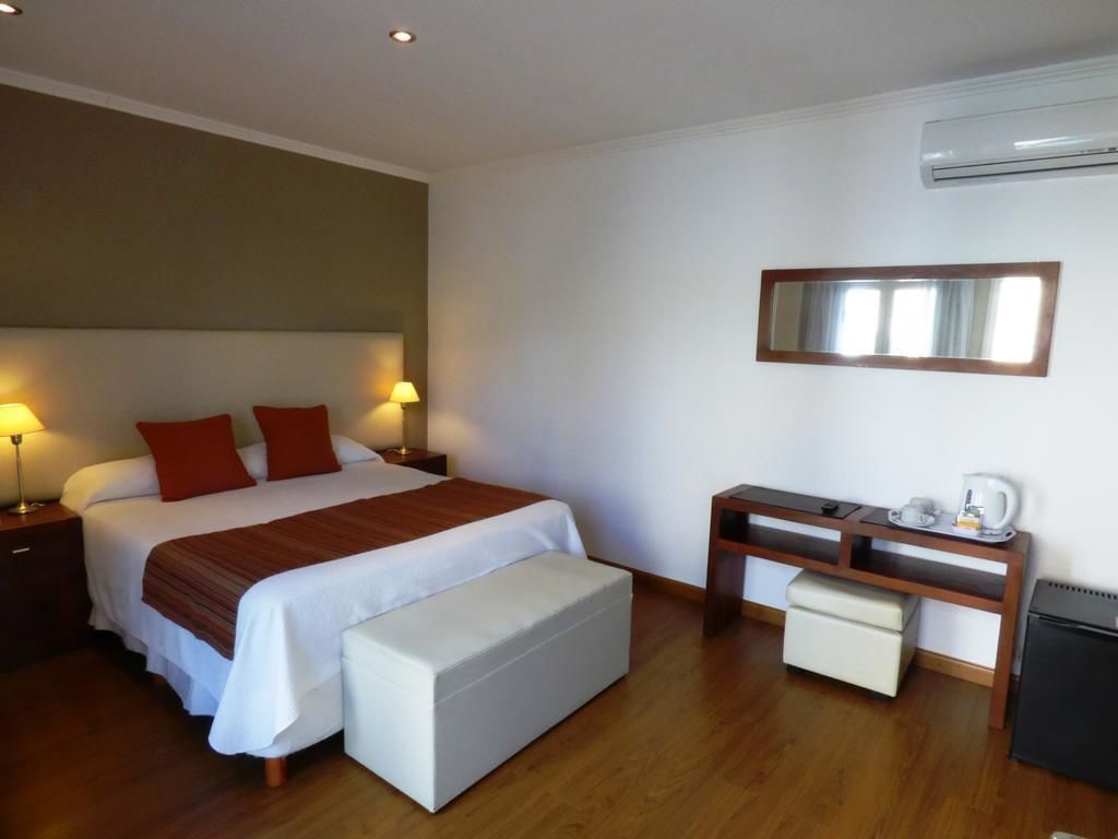 A hotel room with a large bed, bedside lamps, a desk with a chair, a small table with a kettle, and an air conditioning unit.