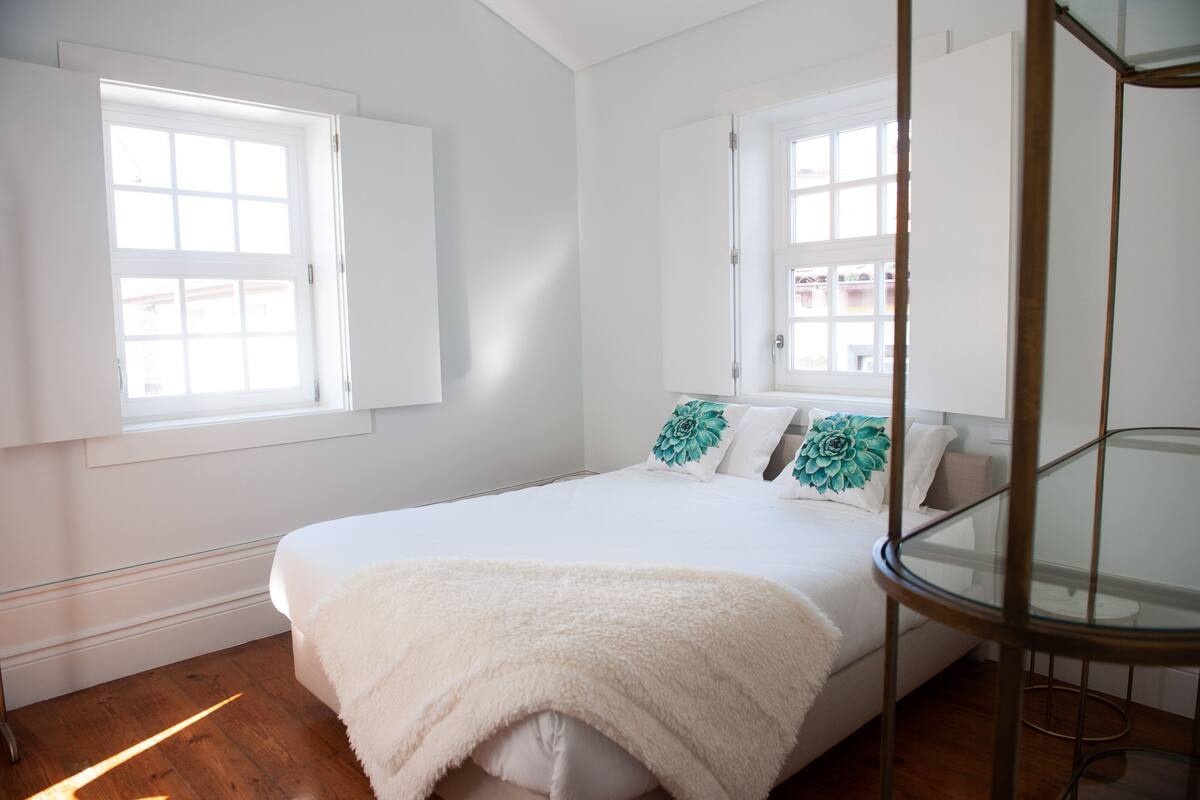 A bedroom with a bed, a glass table, and two windows.