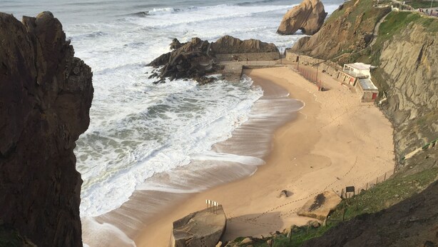 Une plage côtière bordée de falaises rocheuses, avec une étendue de sable et un phare visible au loin.