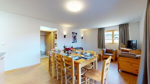 A modern, well-lit dining area with a wooden table set for six, surrounded by chairs. A cozy living area with a sofa and a television is visible in the background. - Pracondu 1 A01 - OUTDOOR & FUN  charming apartment (Haute-Nendaz)