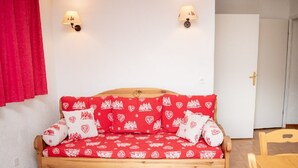 Una habitación acogedora con un sofá cama de madera, dos lámparas de pared, una mesa de madera con vasos y una vela, y una silla de madera.
