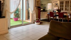 Un salon doté d’une porte-fenêtre coulissante donnant sur un jardin, un coin repas avec une table et des chaises, et une cuisine à l’arrière-plan.