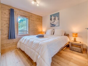 Un dormitorio con suelo de madera, una cama con ropa de cama blanca, una ventana con cortinas, un cuadro en la pared y una mesita de noche con una lámpara.