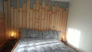 Een slaapkamer met een houten hoofdbord, een bed met een decoratief sprei, twee nachtkastjes met kaarsen en een raam aan de rechterkant.