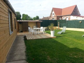 A backyard with a wooden deck, white chairs, a table, and a green lawn.