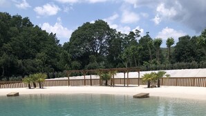 Ein klares Schwimmbecken mit Holzterrasse, umgeben von Sand und Bäumen.