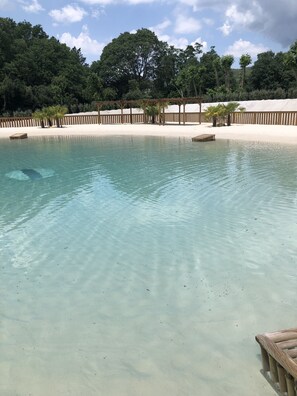 Ein klares Schwimmbecken mit Holzterrasse, umgeben von Sand und Bäumen.