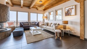 Un salon spacieux doté d’une cheminée en pierre, de poutres apparentes en bois et de grandes fenêtres. On y trouve deux canapés, une table basse et une petite table d’appoint.