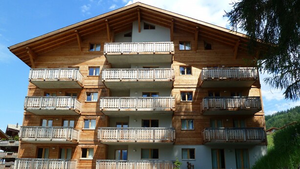 Un immeuble en bois à plusieurs étages, doté de balcons, d’une entrée centrale et entouré d’un paysage naturel.