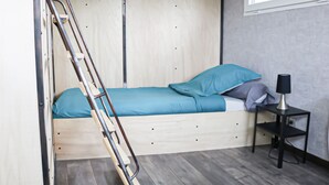 A small, compact room with a bunk bed, a ladder, a small table with a lamp, and a window providing natural light. - Bay Terminus - The Hanging Lodge (Saint-Blimont)