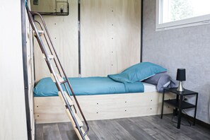 A small, compact room with a bunk bed, a ladder, a small table with a lamp, and a window providing natural light.