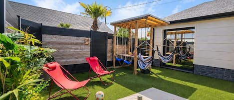 Un jardin avec une pelouse artificielle, deux fauteuils de jardin rouges, un hamac et une cabane en bois.