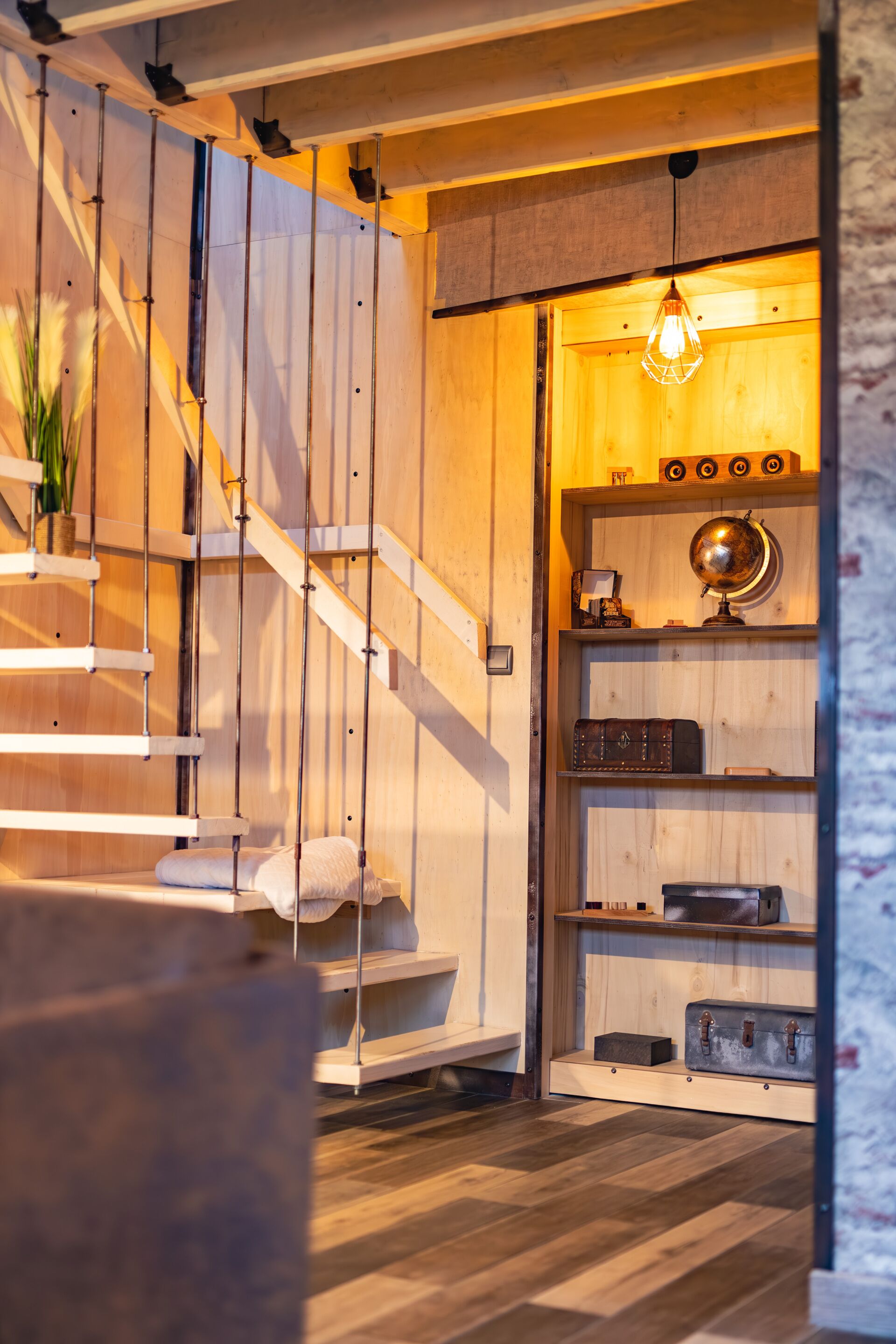 Ein Holzinterieur mit Treppe, einem Bett, einem Regal mit Büchern und einem Globus, sowie einer hängenden Leuchte.