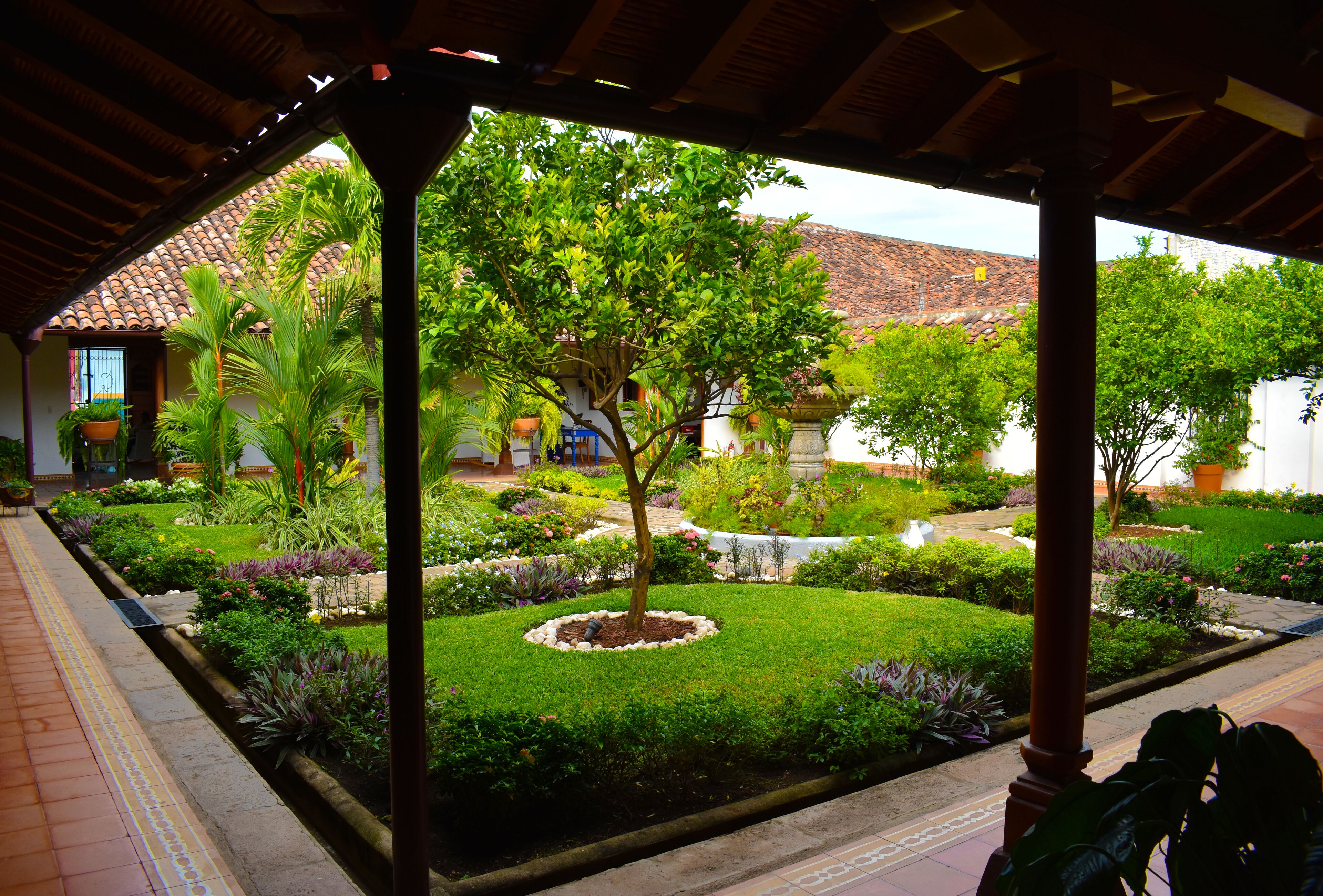 Un patio con techo de tejas, un jardín con diversas plantas y un edificio con techo rojo al fondo.