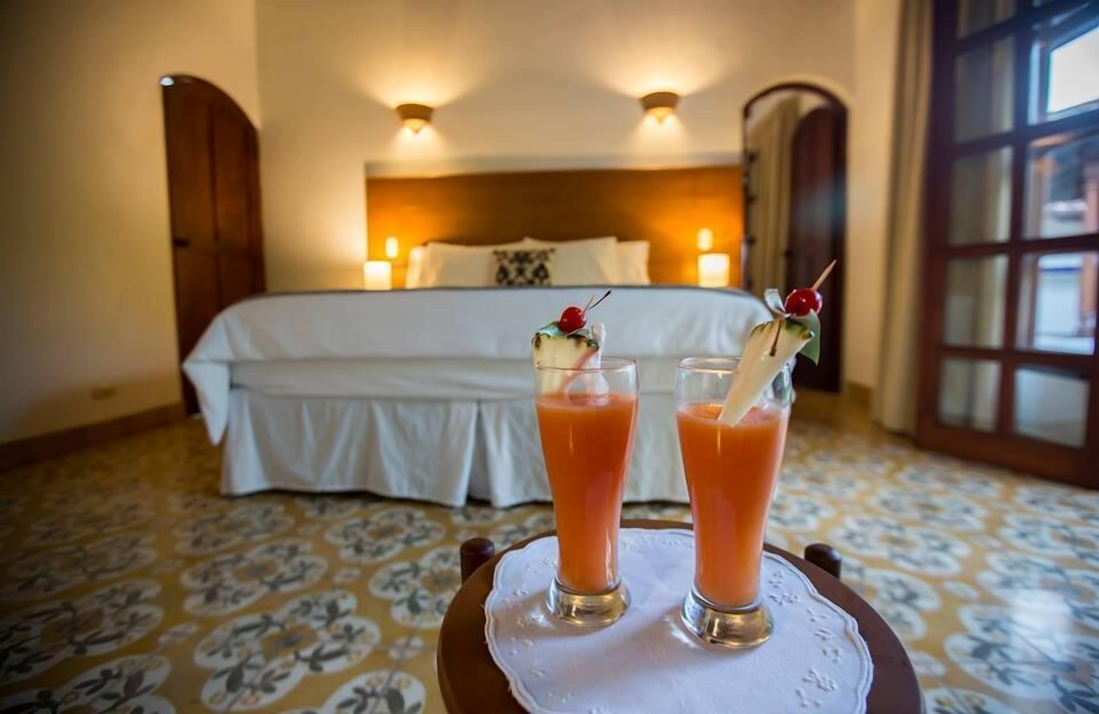 Une chambre d’hôtel avec un lit soigneusement fait, deux verres de jus d’orange sur une table et une grande fenêtre avec des rideaux.