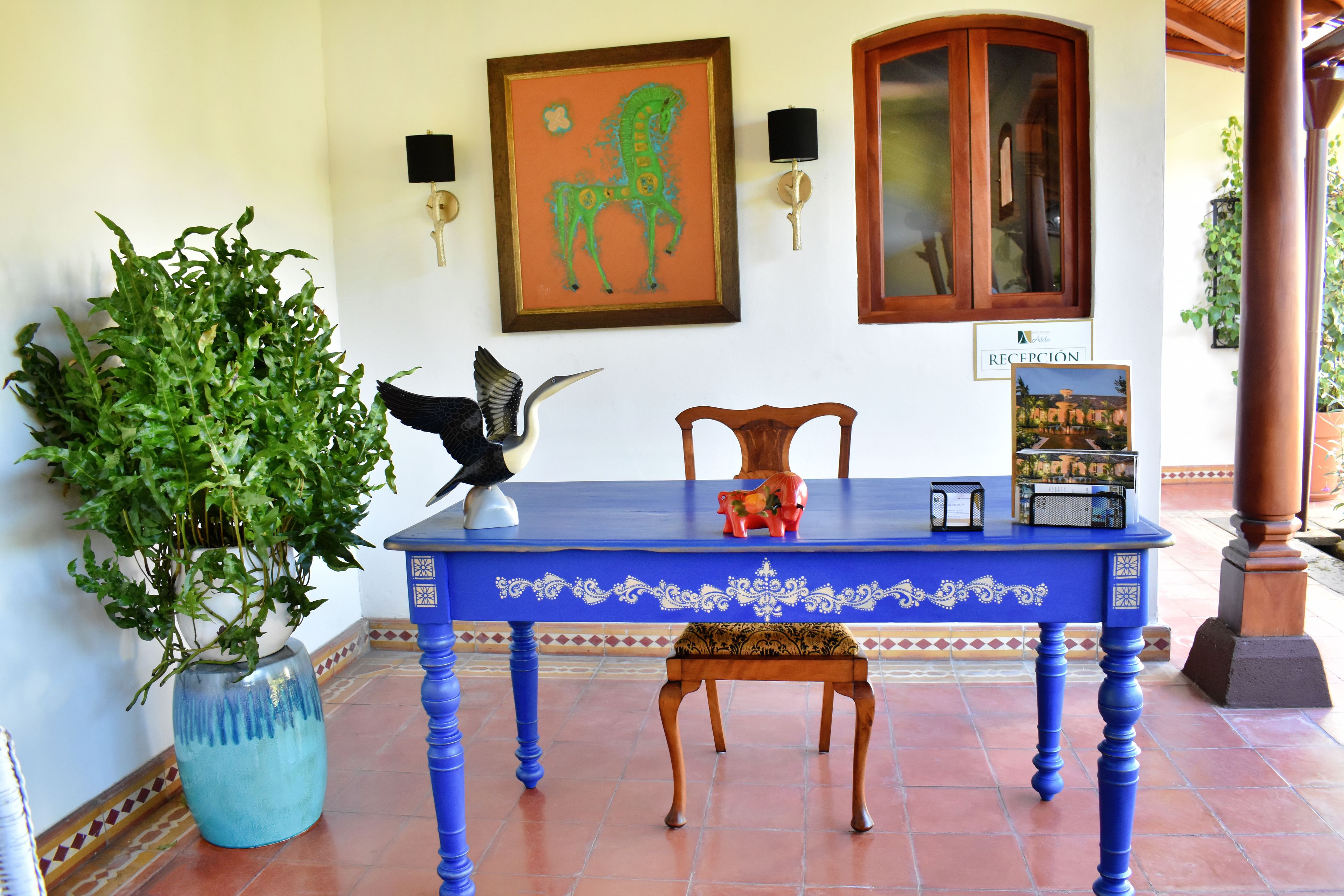 A reception area with a blue table, a wooden chair, a potted plant, a framed horse painting, and a cabinet.