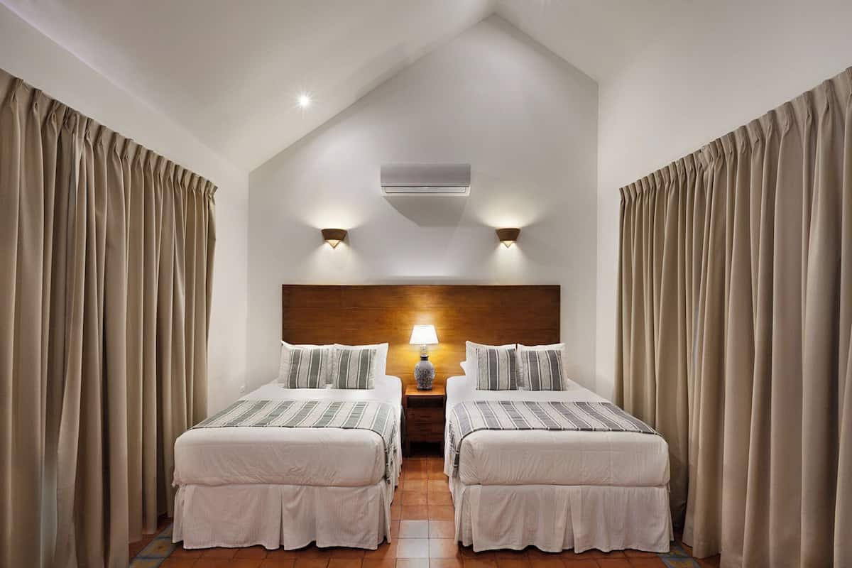 A hotel room with two beds, beige curtains, a wooden headboard, a nightstand with a lamp, and an air conditioning unit.