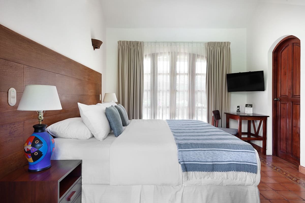 A hotel room with a wooden headboard, a bed with white bedding and a striped blanket, a desk with a lamp, a TV, and a door.