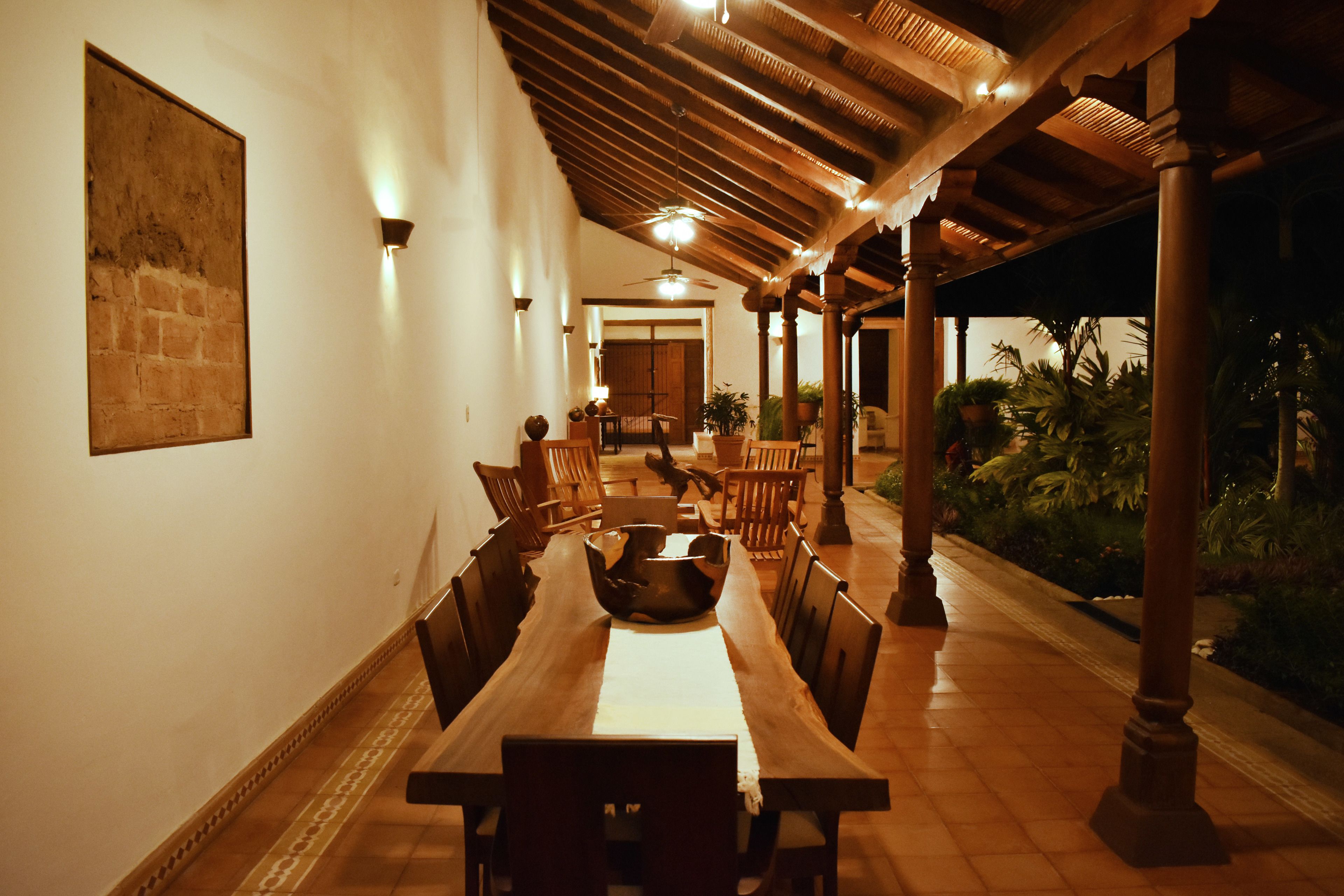 Un área de comedor techada con una larga mesa de madera y sillas, un ventilador de techo y plantas en macetas.