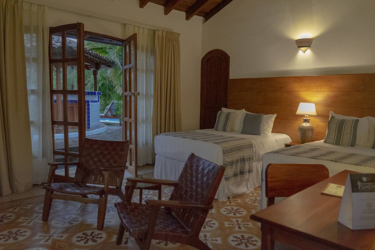A hotel room with two beds, a wooden headboard, a table with a lamp, and a chair.