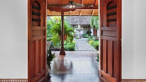 A wooden door leading to a tiled patio with a ceiling fan, surrounded by greenery and a stone pathway.