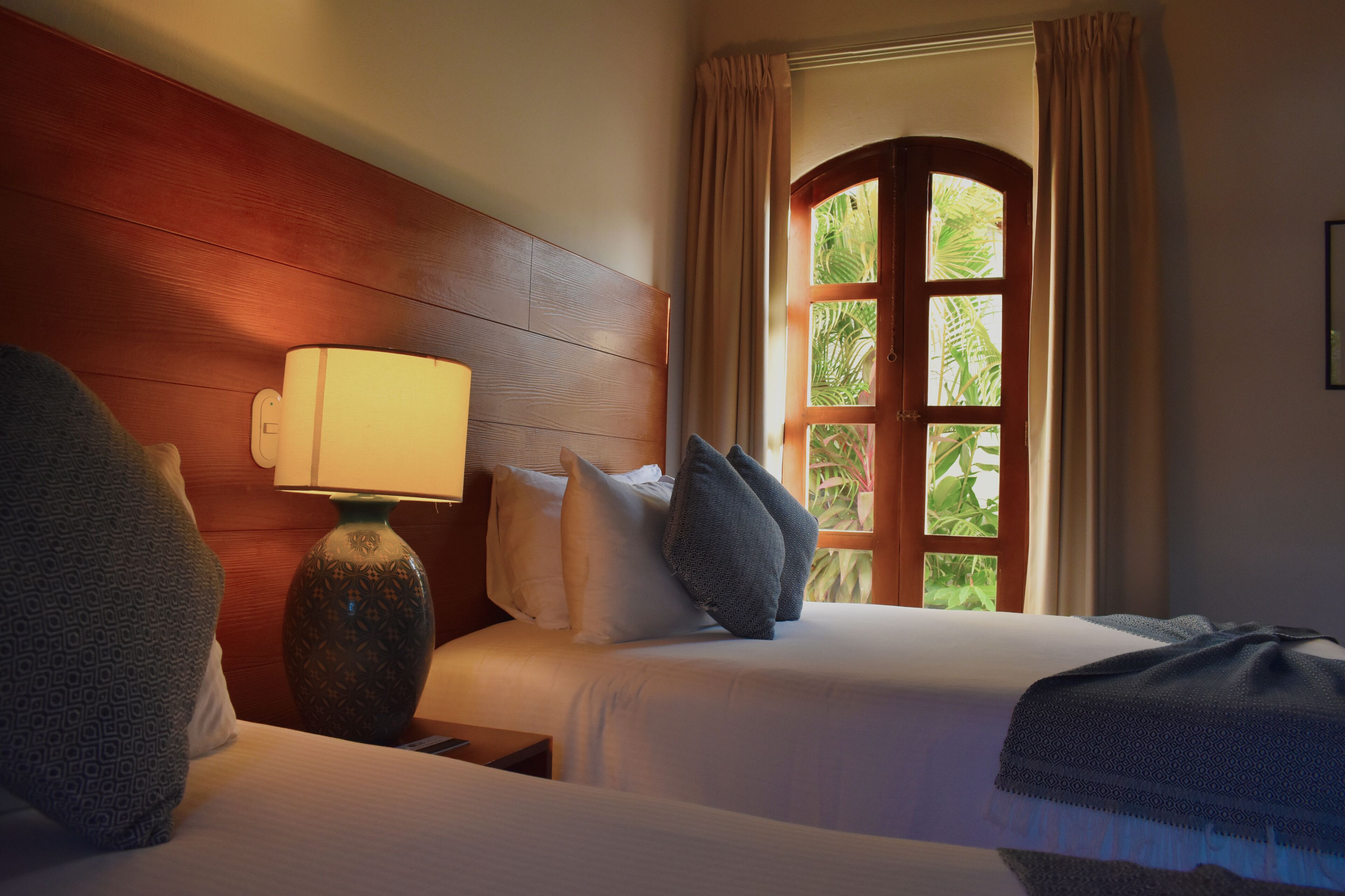 Habitación de hotel con dos camas, cabecera de madera, lámpara de noche y ventana con cortinas.