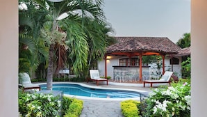 A pool area with a wooden pergola, lounge chairs, and a small building with a tiled roof.