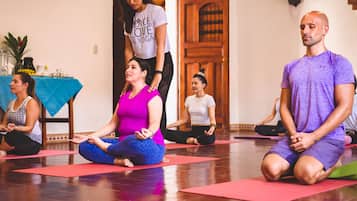 Eine Gruppe von Menschen übt Yoga auf roten Matten in einem Raum mit Holzdielen und einer blauen Tischdecke.