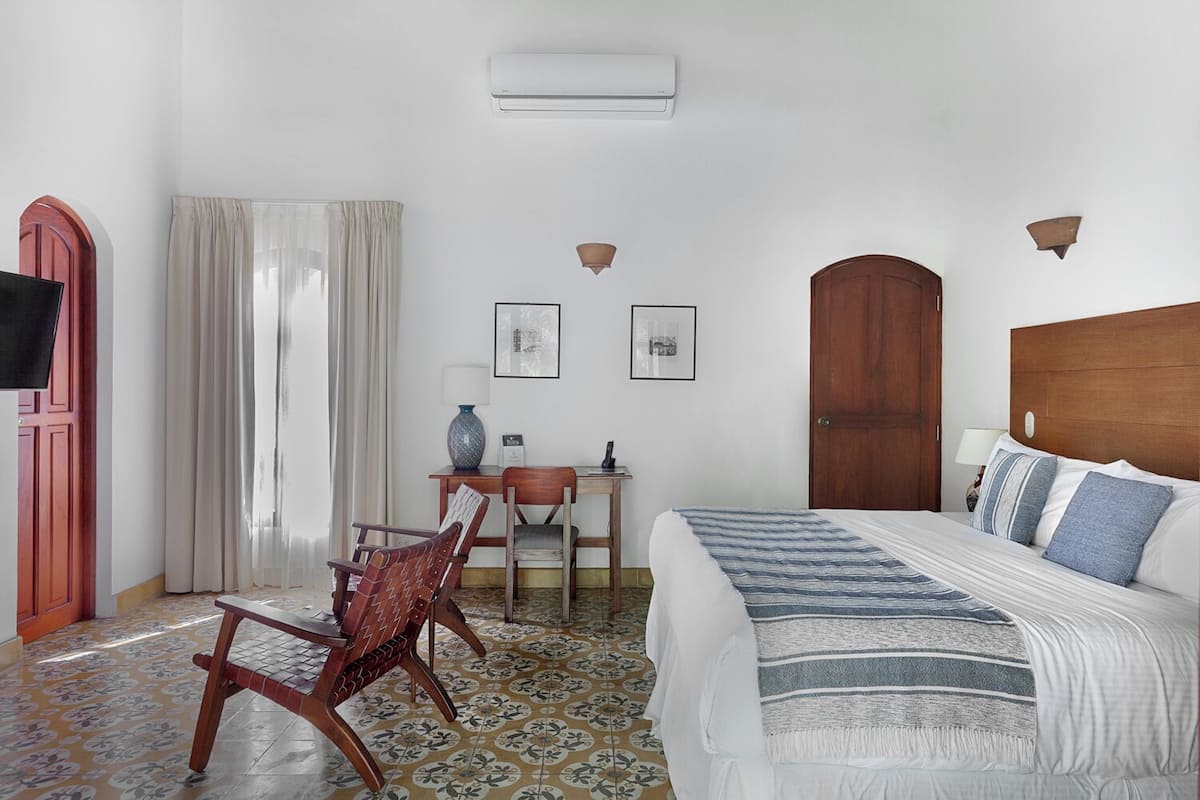 A hotel room with a bed, a desk, a chair, a wooden door, and a wall-mounted air conditioner.
